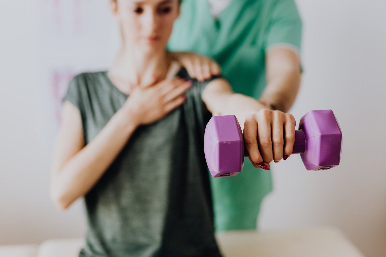 Crop anonymous osteopath assisting patient with dumbbell in hand